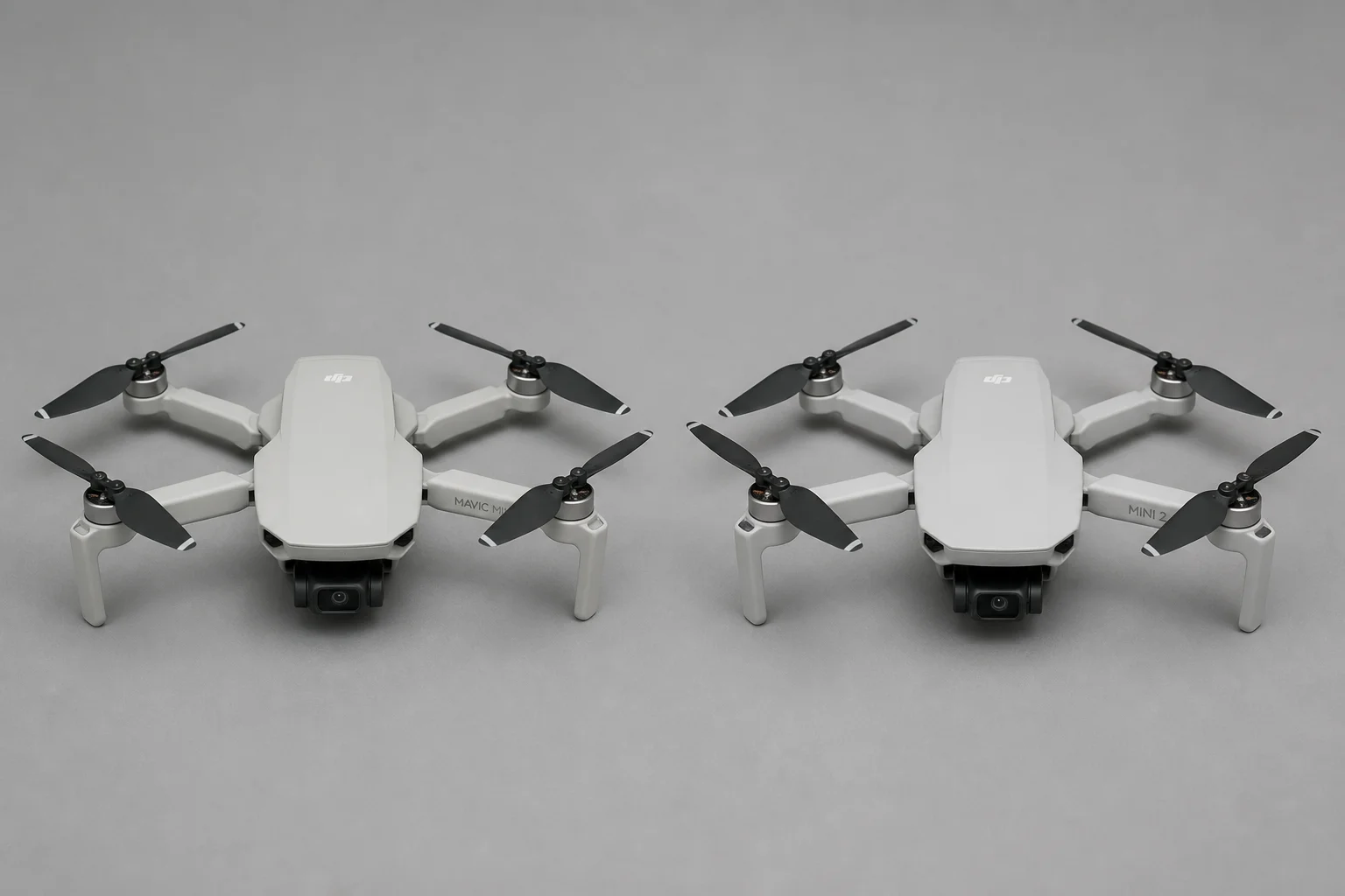 DJI Mavic Mini and DJI Mini 2 placed side by side on a flat surface, showing their similar compact folded-arm design